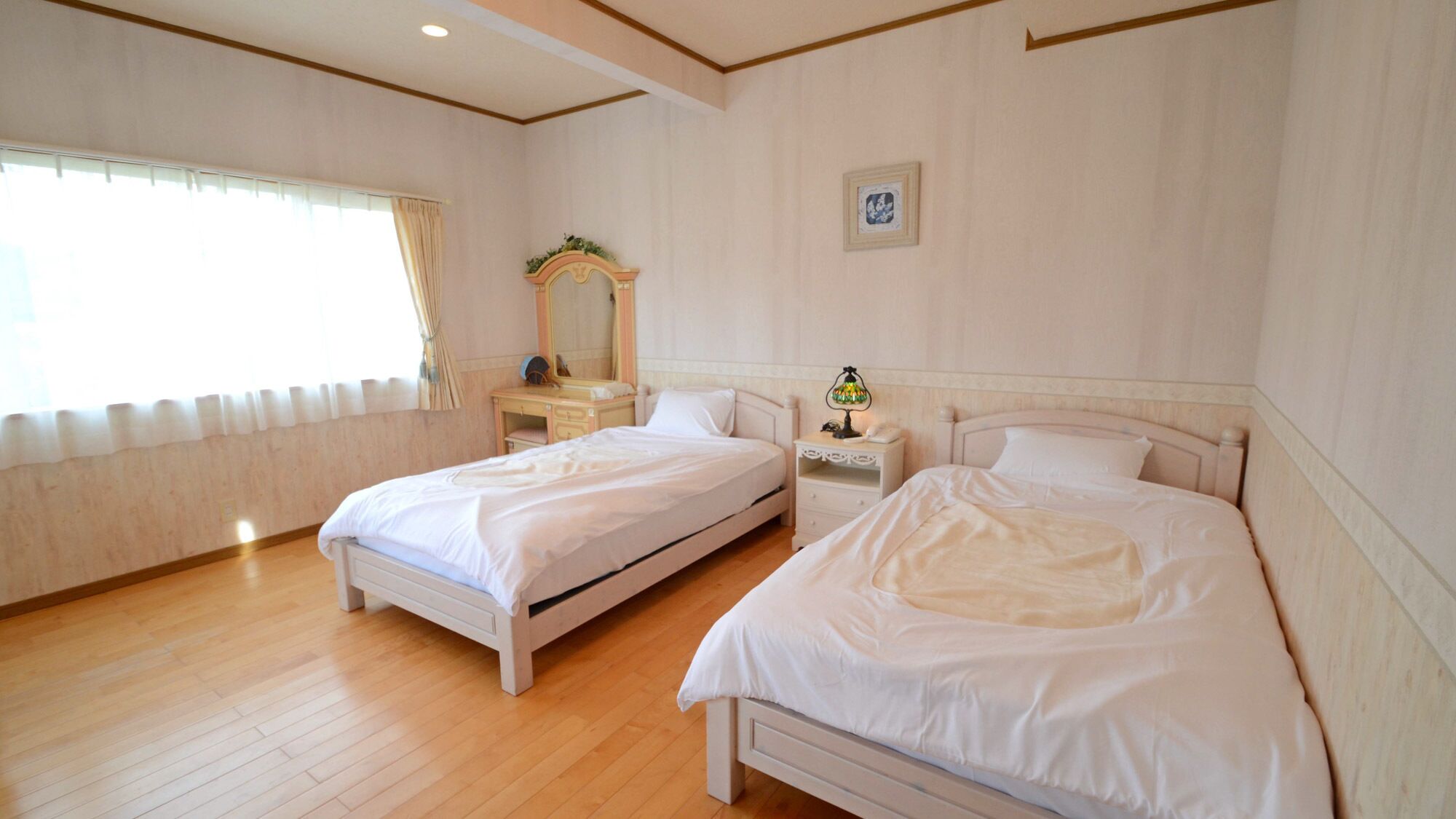 A hotel room with two beds, a vanity with a mirror, a bedside table with a lamp, and a framed picture on the wall.