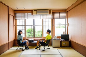 Una habitación japonesa tradicional con suelo de tatami, un televisor sobre un mueble y vistas a la vegetación exterior.
