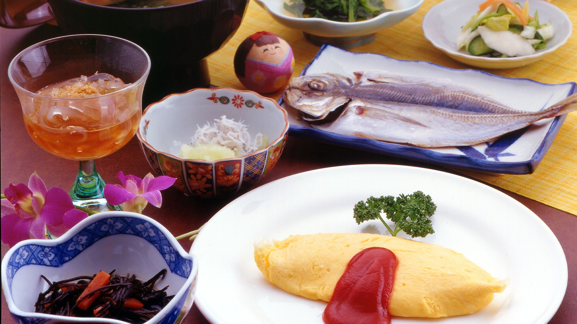 A table with various dishes including fish, a bowl of shredded vegetables, a glass of tea, and a plate with an omelette and a red garnish.