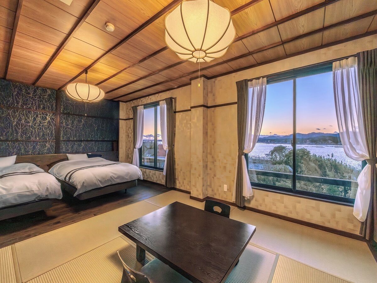 Una habitación con cama, una mesa y ventanas grandes con cortinas.