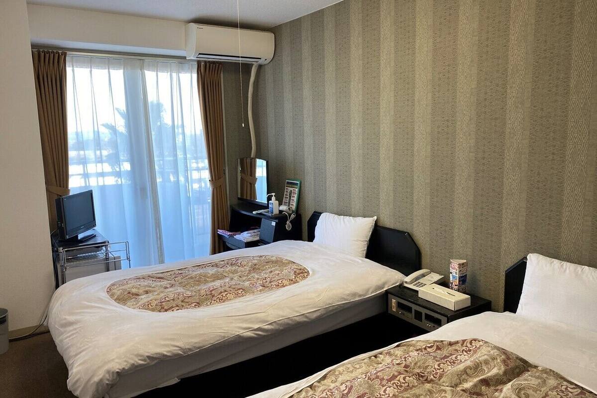 A hotel room with two beds, a desk with a computer, a telephone, and a window with curtains.