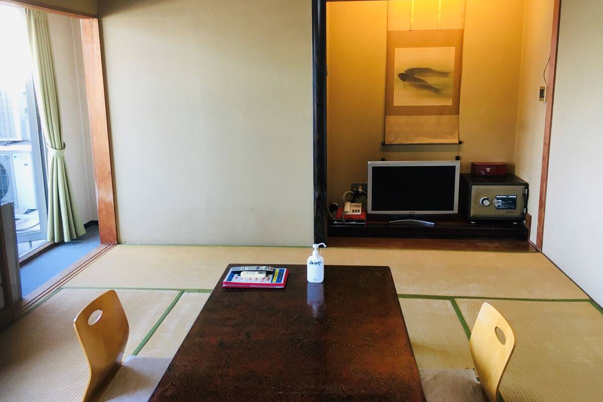 A traditional Japanese room with tatami flooring, a low table, two chairs, a television, and a safe.