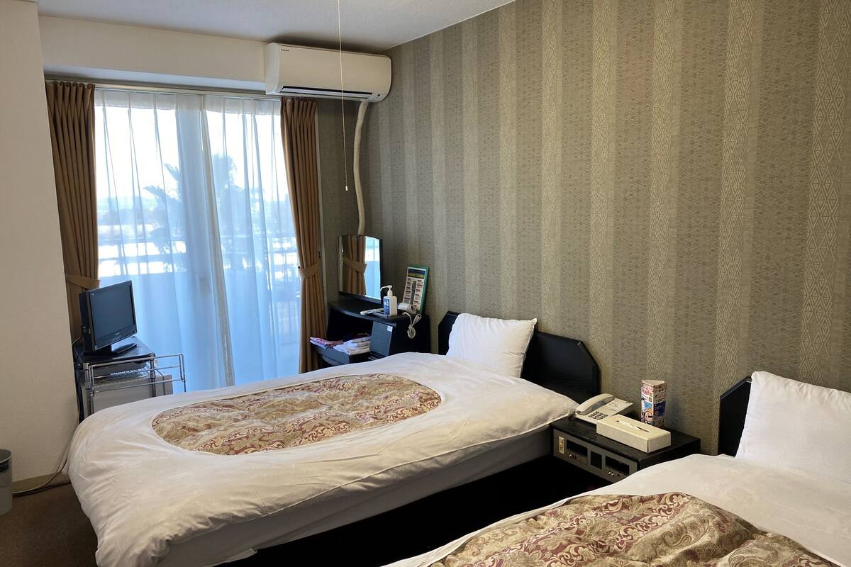 A hotel room with two beds, a desk with a computer, a telephone, and an air conditioning unit.