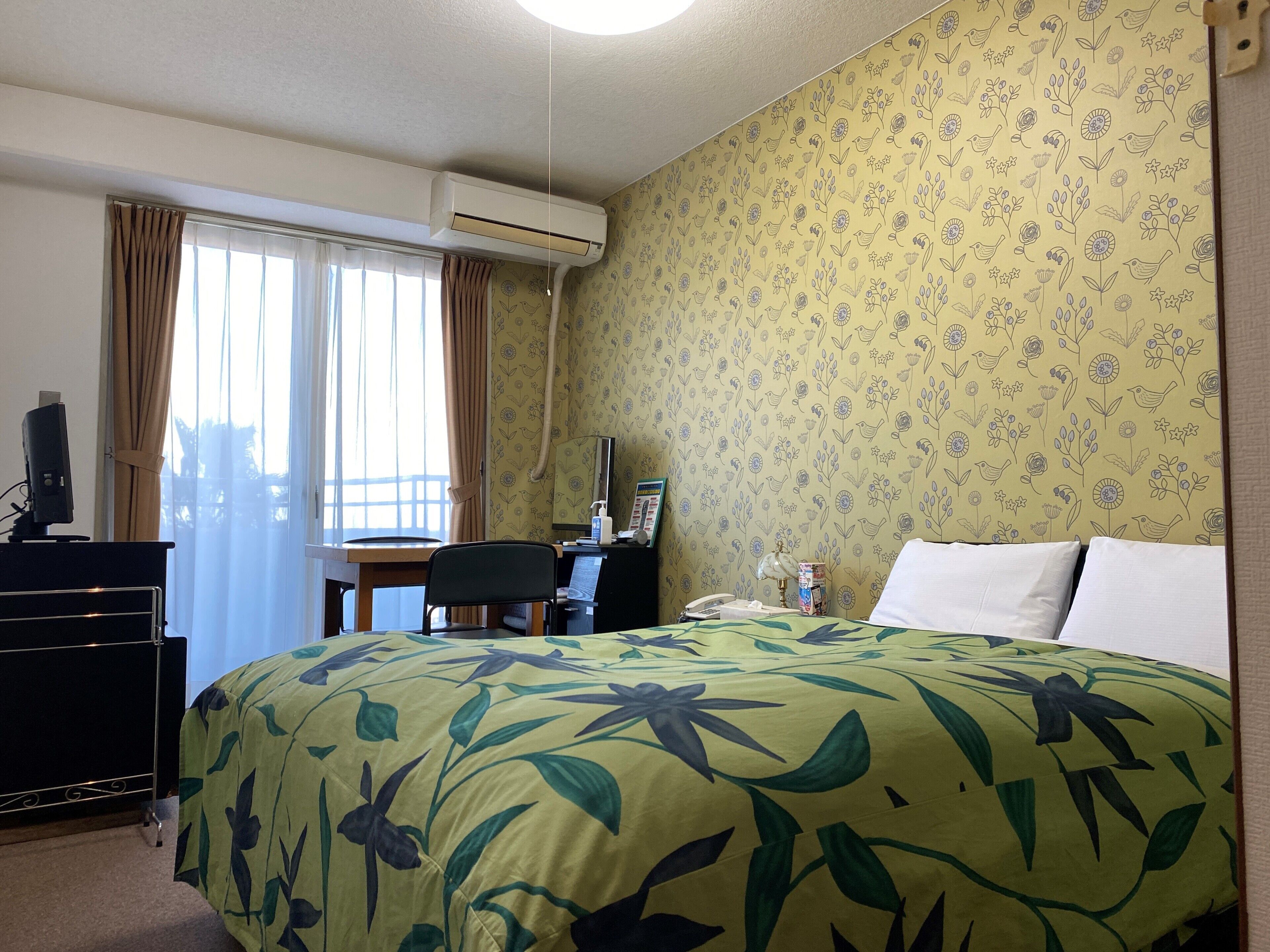 A hotel room with a bed, desk, chair, air conditioning unit, and a window with curtains.