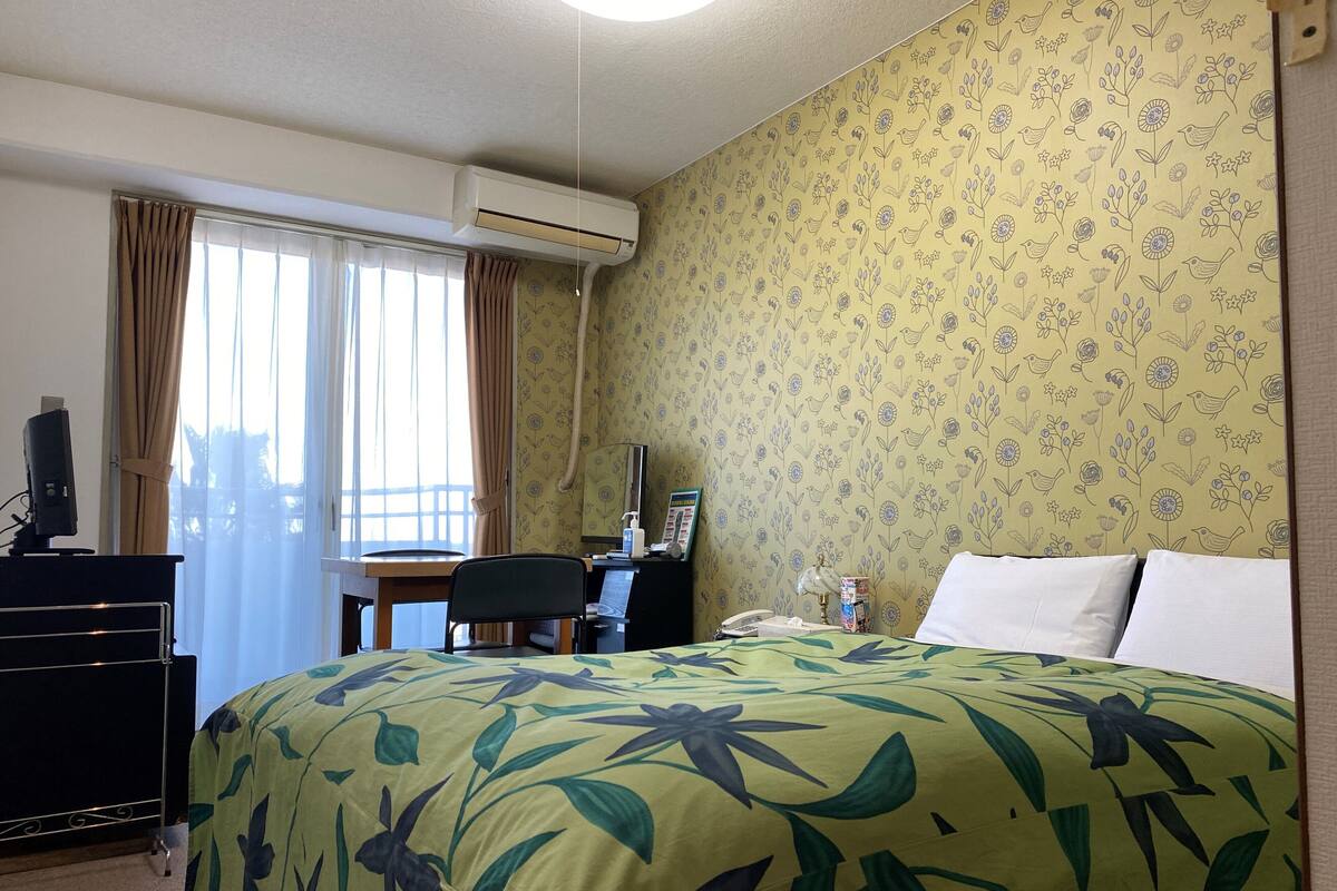 A hotel room with a bed, desk, chair, air conditioning unit, and a window with curtains.