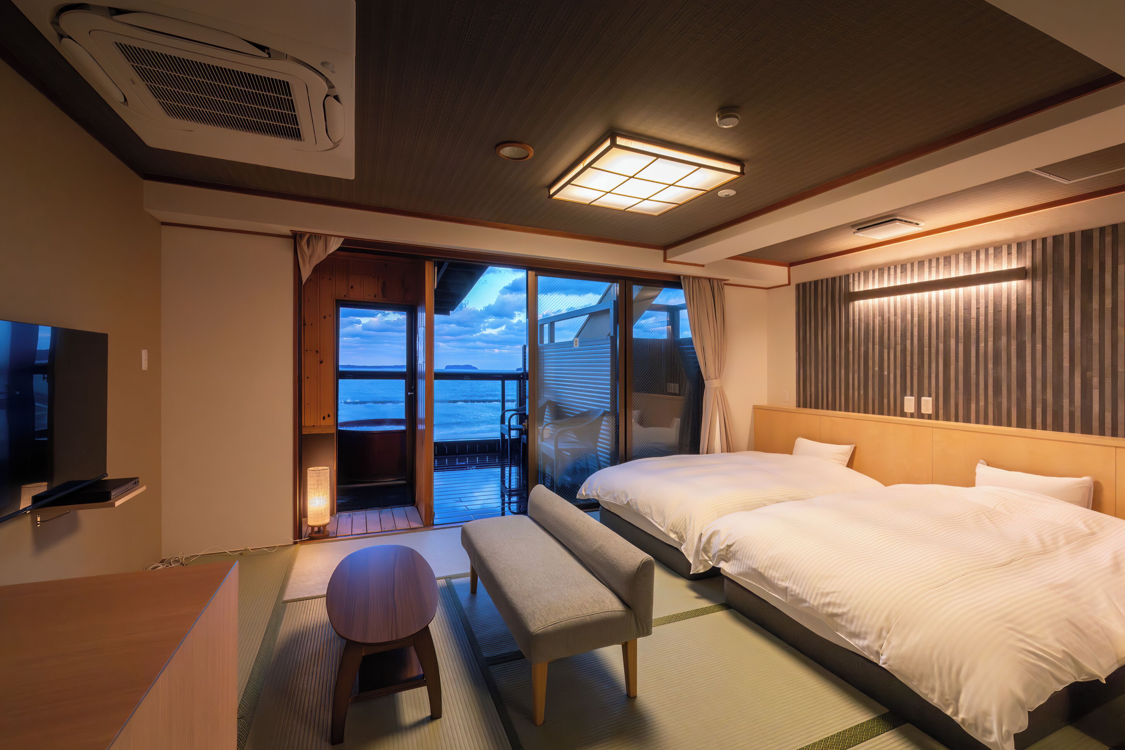 Habitación de hotel con cama, un banco, televisor y vista al mar.