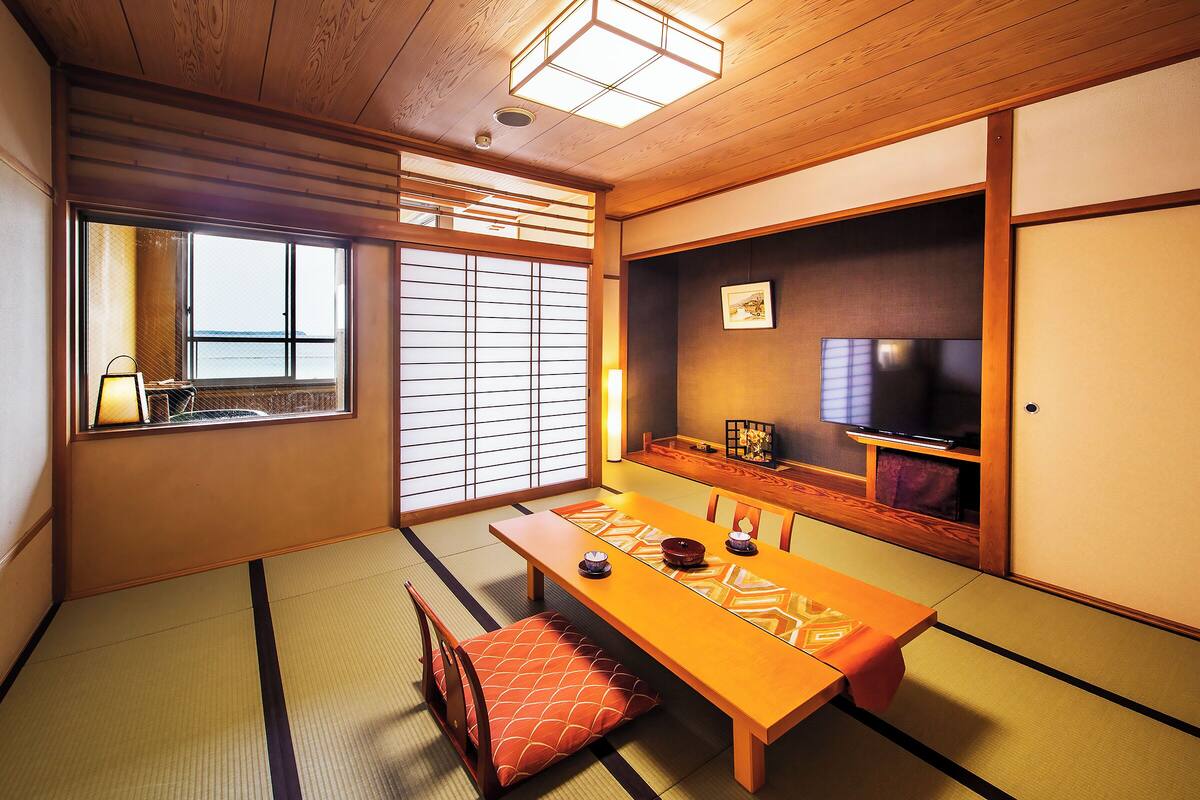 A traditional Japanese room with tatami flooring, a low table, cushions, a sliding door, and a television.