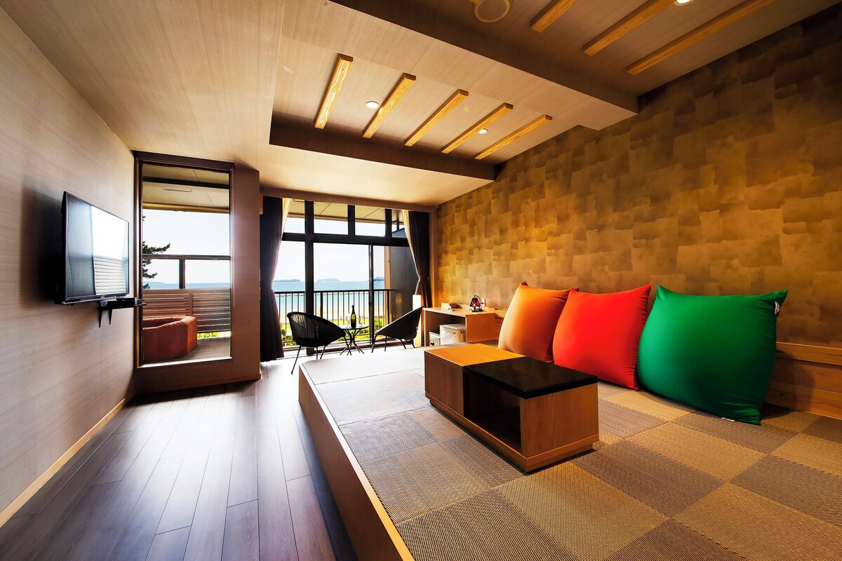 A modern living room with a sofa, a coffee table, and a TV mounted on the wall. There is a balcony with a view of the sea.
