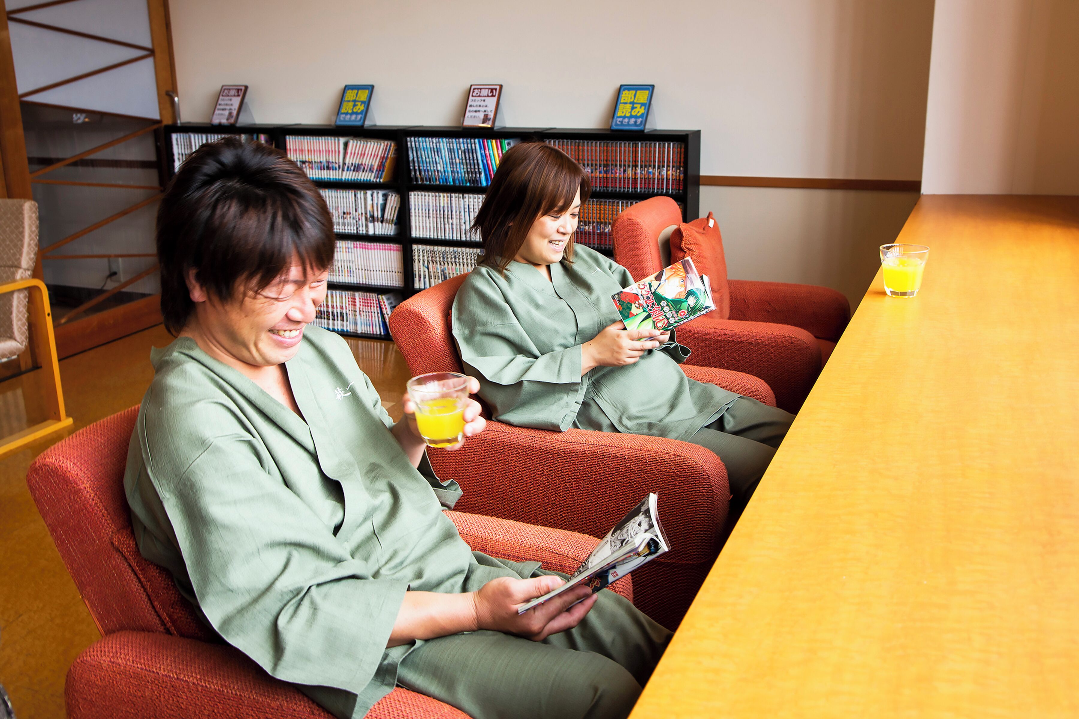 Two people sitting on red chairs at a wooden table with a glass of orange juice, reading magazines in a room with a bookshelf in the background.