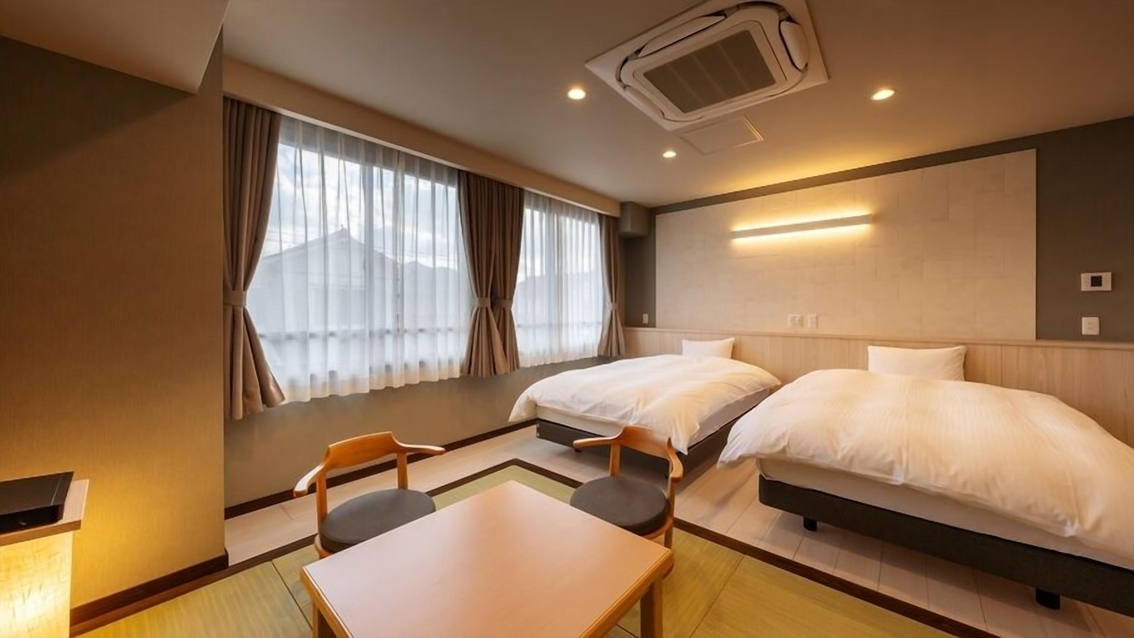 A hotel room with two beds, a table, chairs, a window with curtains, and an air conditioning unit.