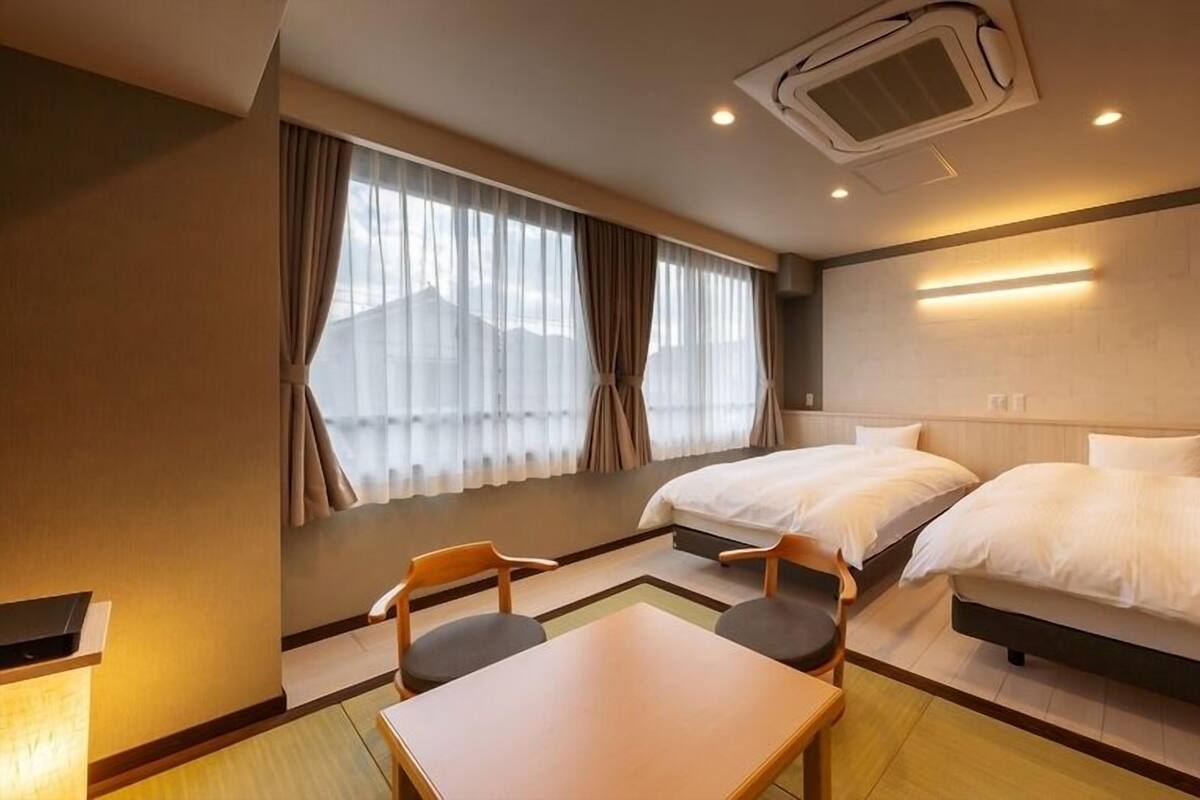 A hotel room with two beds, a table, chairs, a window with curtains, and an air conditioning unit.