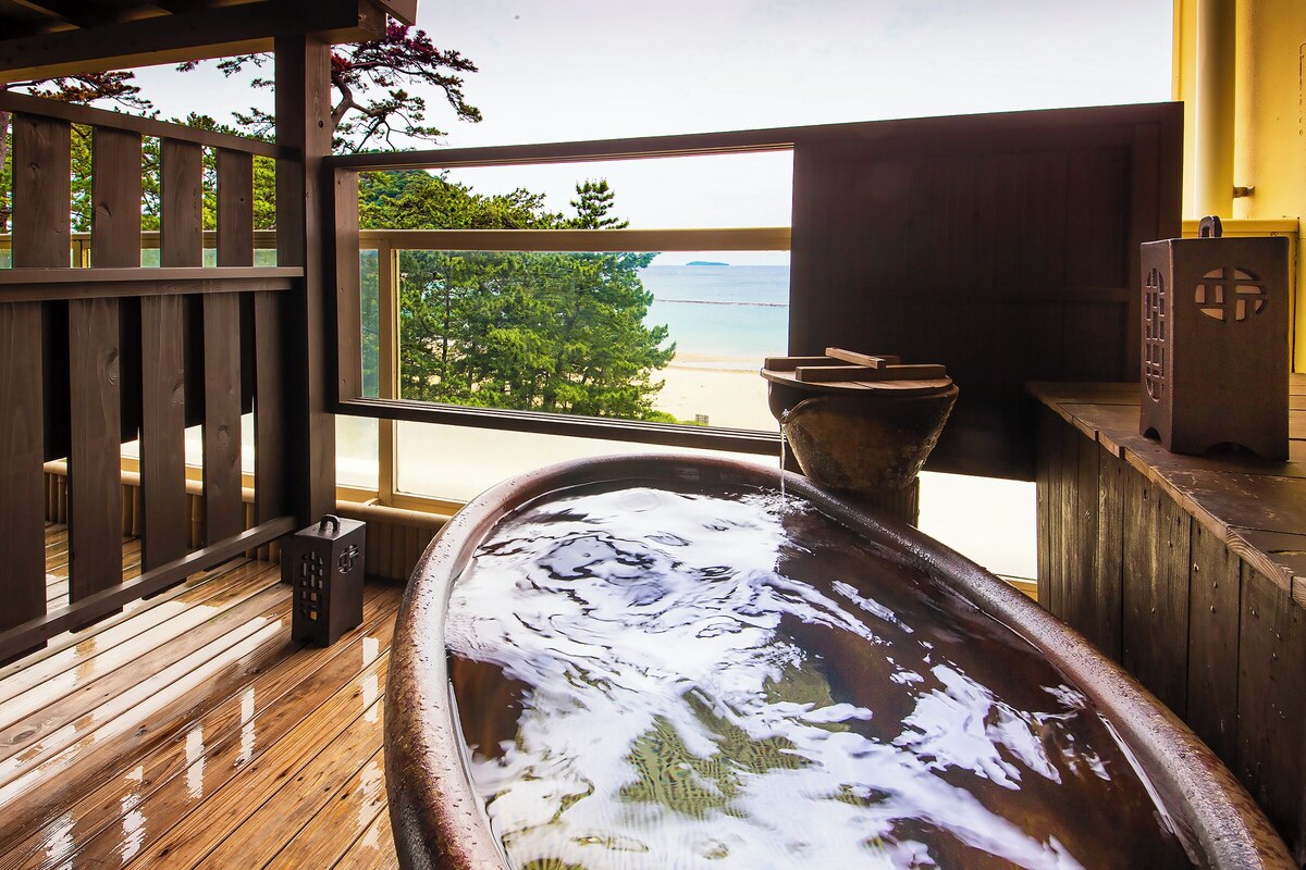 A wooden deck with a large bathtub, a view of the beach, and a wooden fence.