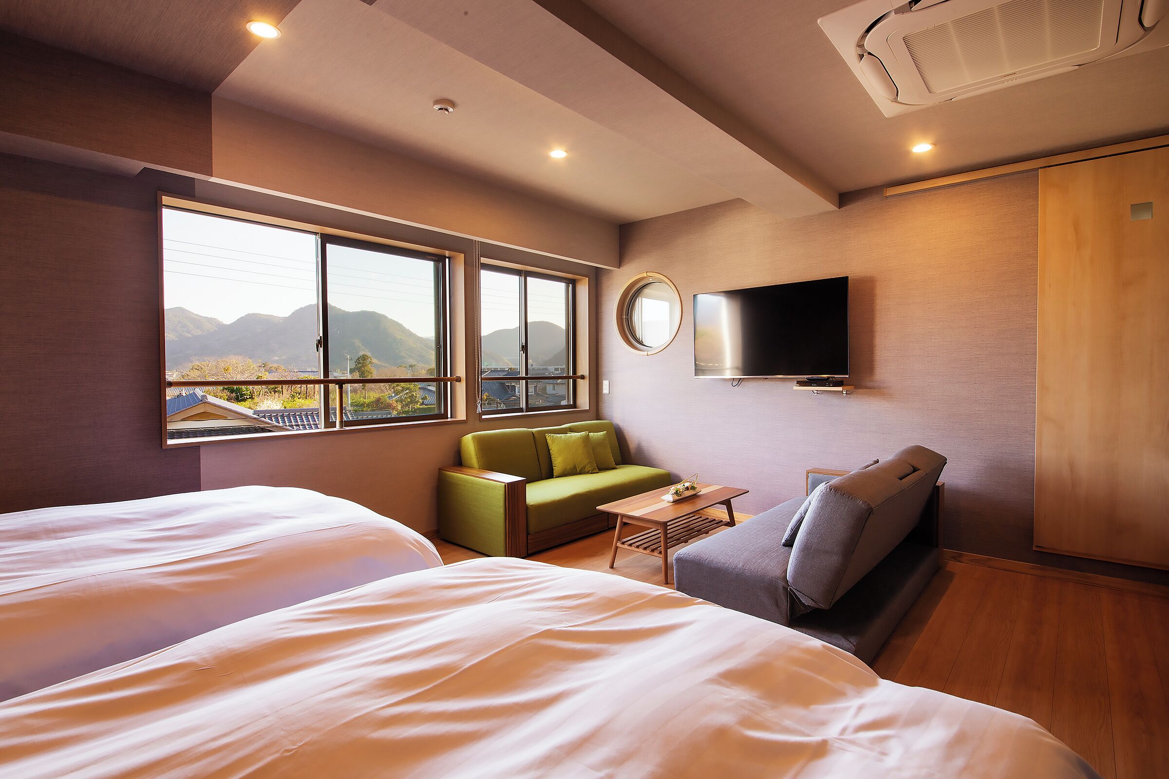 A hotel room with two beds, a sofa, a small table, a TV, and a large window offering a mountain view.