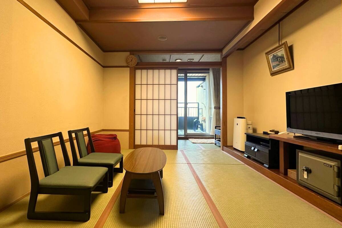 A traditional Japanese-style room with tatami flooring, a low table with cushions, a television, and a sliding door leading to a balcony.
