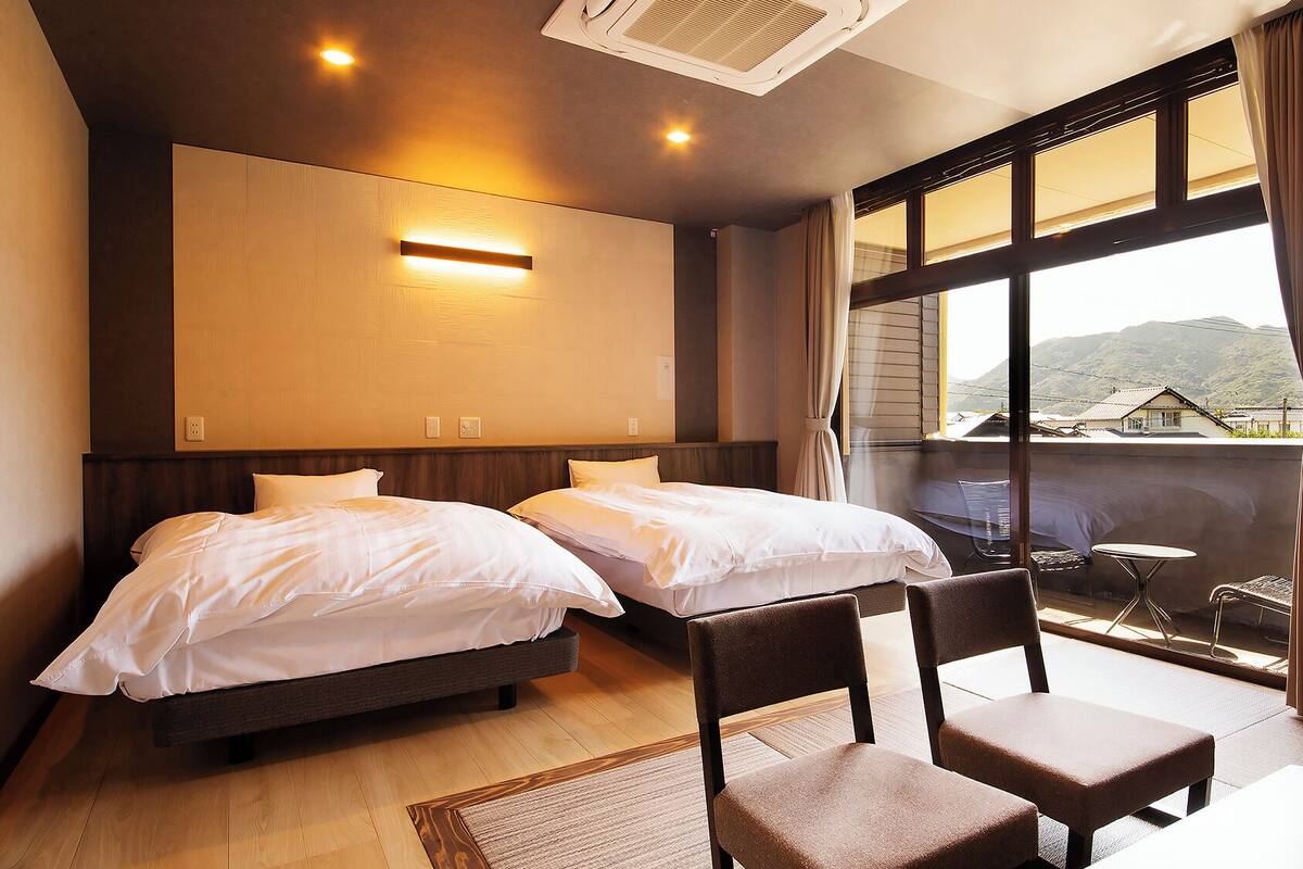 A hotel room with two beds, a chair, a small table, a balcony with a view of mountains, and a ceiling light.