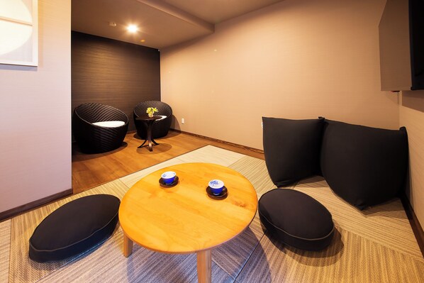 A modern room with a wooden table, two black chairs, a small round table with two blue cups, and black cushions on the floor. - YOIMACHINOYADO HAGIICHIRIN (Hagi)