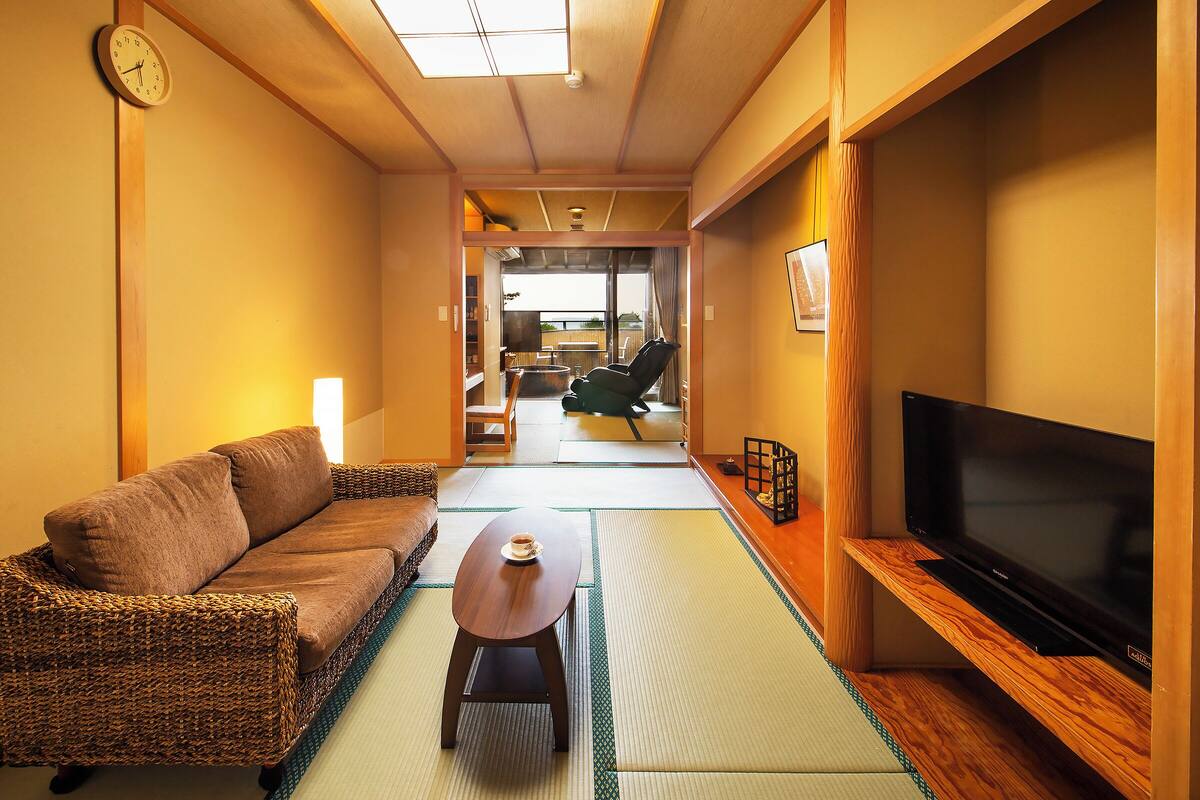 A traditional Japanese-style living room with tatami flooring, a low table with a cup, a sofa, and a television on a wooden stand.