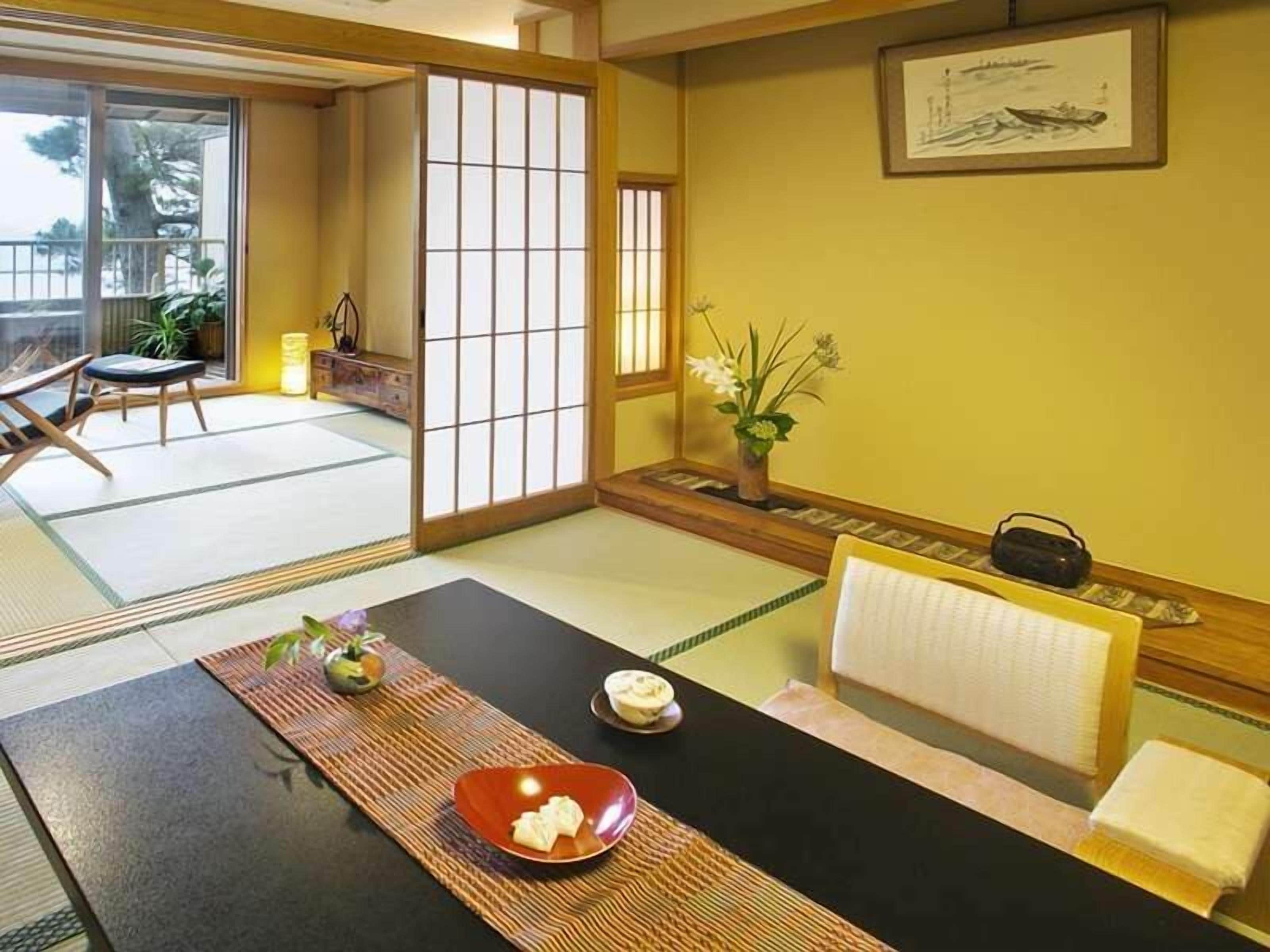 Ett traditionellt japanskt rum med tatamimattor på golvet, ett lågt bord med en röd skål, en liten vas med blommor och en inramad tavla på väggen.