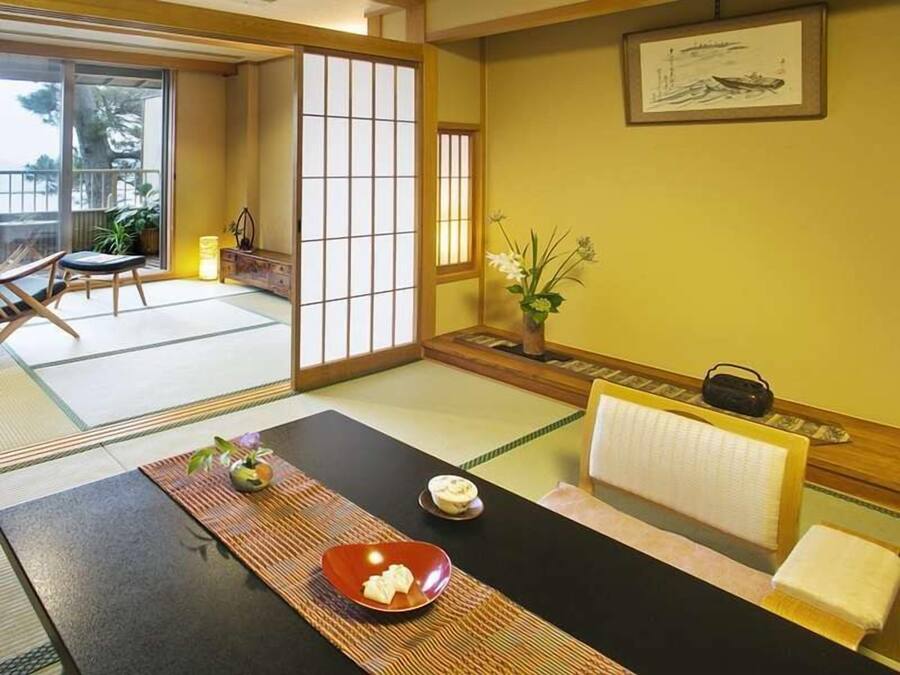 Ett traditionellt japanskt rum med tatamimattor på golvet, ett lågt bord med en röd skål, en liten vas med blommor och en inramad tavla på väggen.