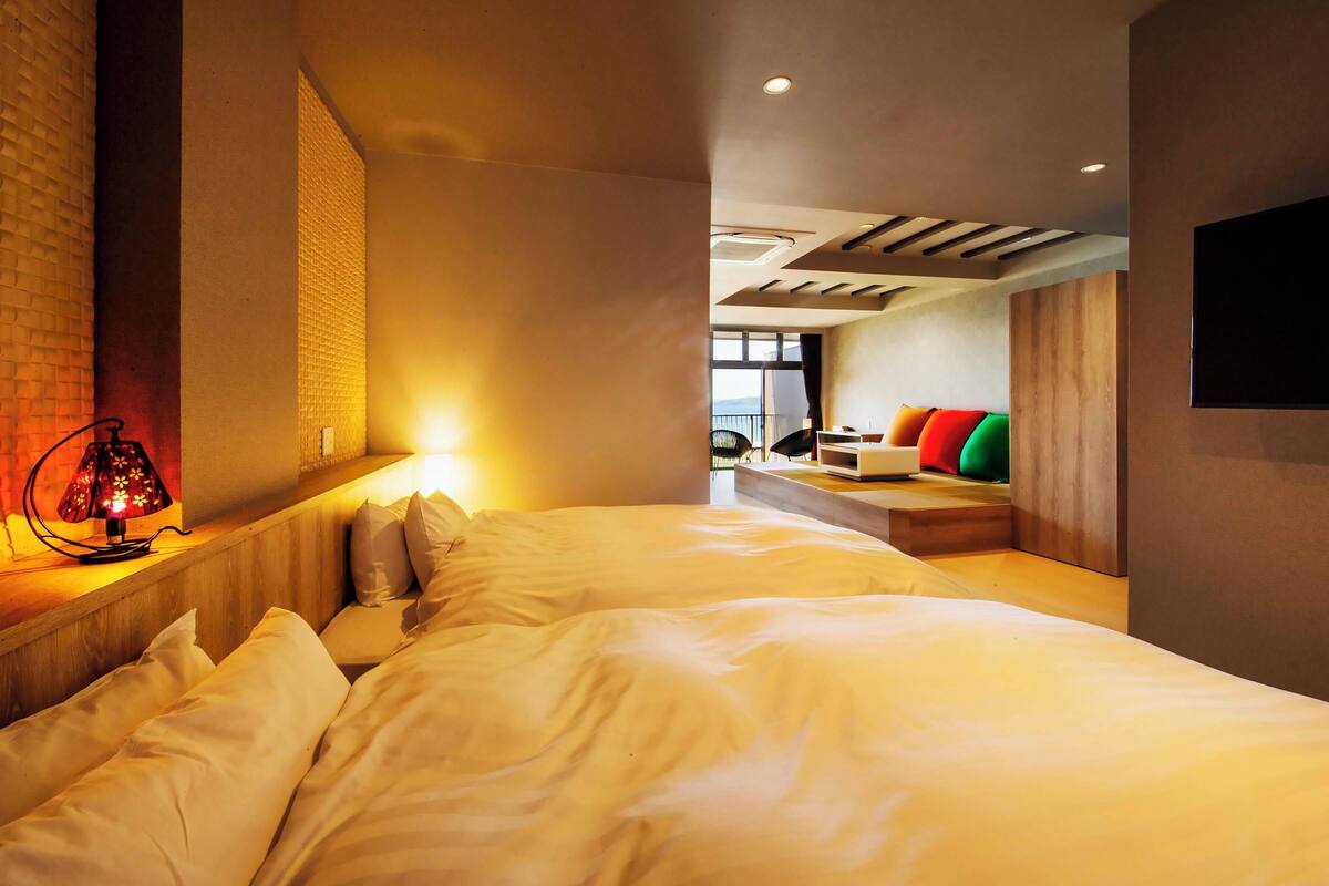 A hotel room with a bed, bedside lamp, and a seating area with colorful cushions.