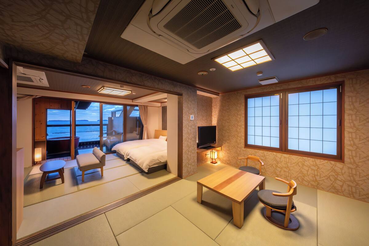 A hotel room with tatami flooring, a bed, a television, a table, and a view of the ocean.