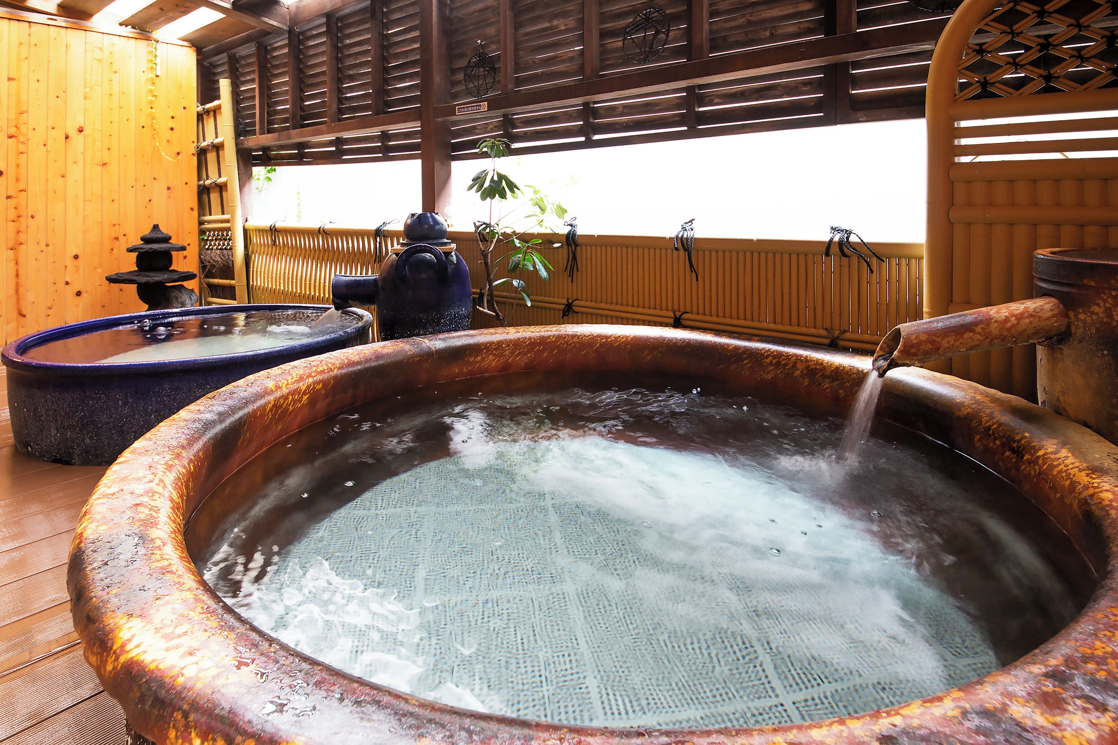 A hot spring area with a large copper hot tub and a smaller one, surrounded by wooden structures and bamboo decorations.