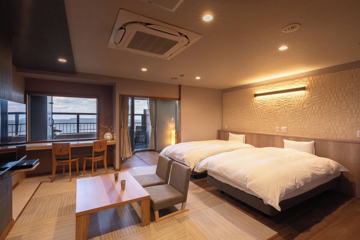 A hotel room with a bed, a desk, chairs, a window with a view, and a brick-patterned wall.