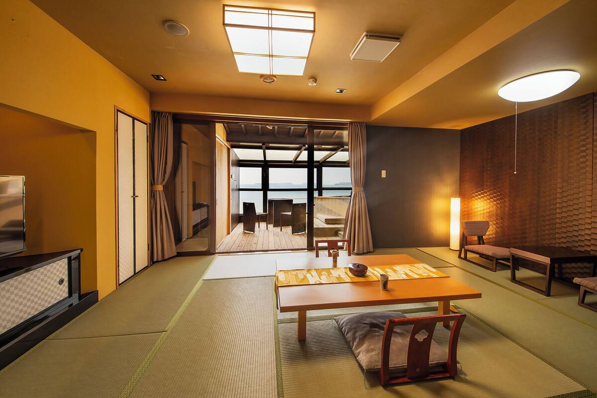 A room with tatami flooring, a low table, a sofa, and a view through a large window.