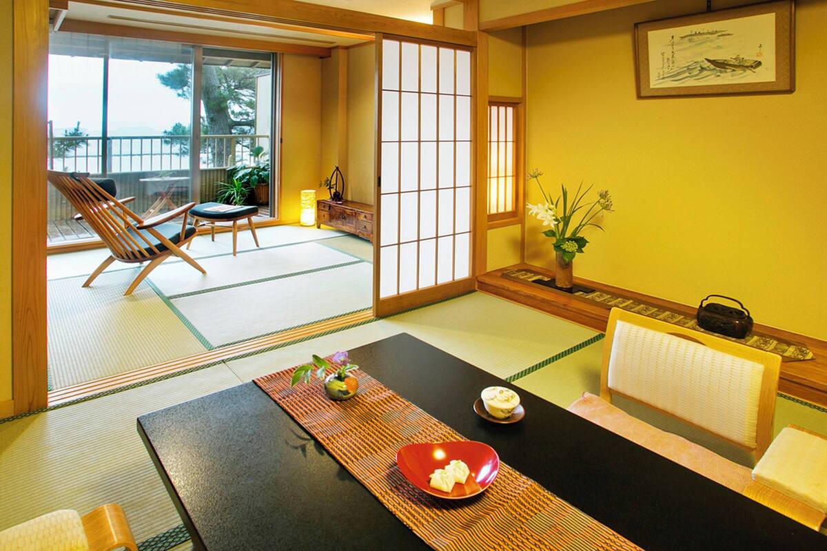 A traditional Japanese room with tatami flooring, a low table with a red plate, a wooden chair, and a sliding door leading to a balcony.