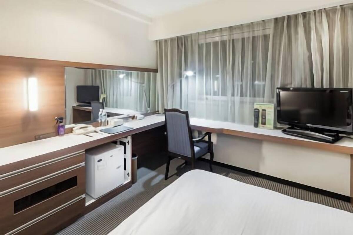 Une chambre d’hôtel comprenant un lit, un bureau avec un ordinateur, une télévision, une chaise et un petit réfrigérateur.