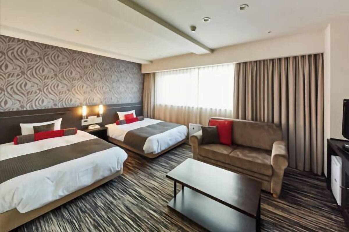 Une chambre d’hôtel avec deux lits, un canapé, une télévision et une grande fenêtre avec des rideaux.