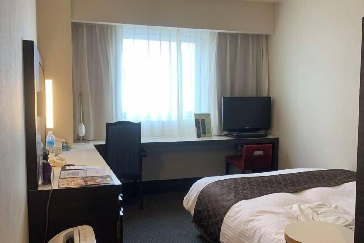 Une chambre d’hôtel comprenant un lit, un bureau avec une chaise, une télévision et une fenêtre avec des rideaux.