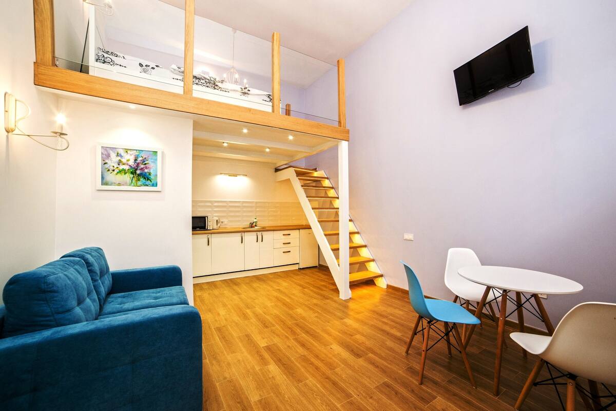Un appartement moderne avec une chambre en mezzanine, un coin repas avec une table blanche et des chaises bleues, une cuisine avec des meubles blancs et un escalier avec des rampes en bois.