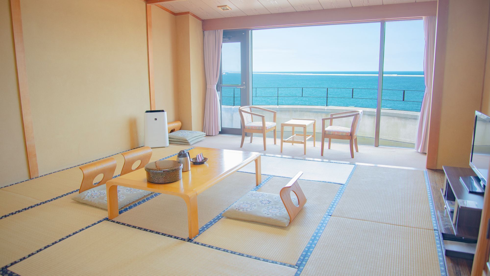 A room with tatami flooring, a wooden table, chairs, a TV, and a view of the sea.
