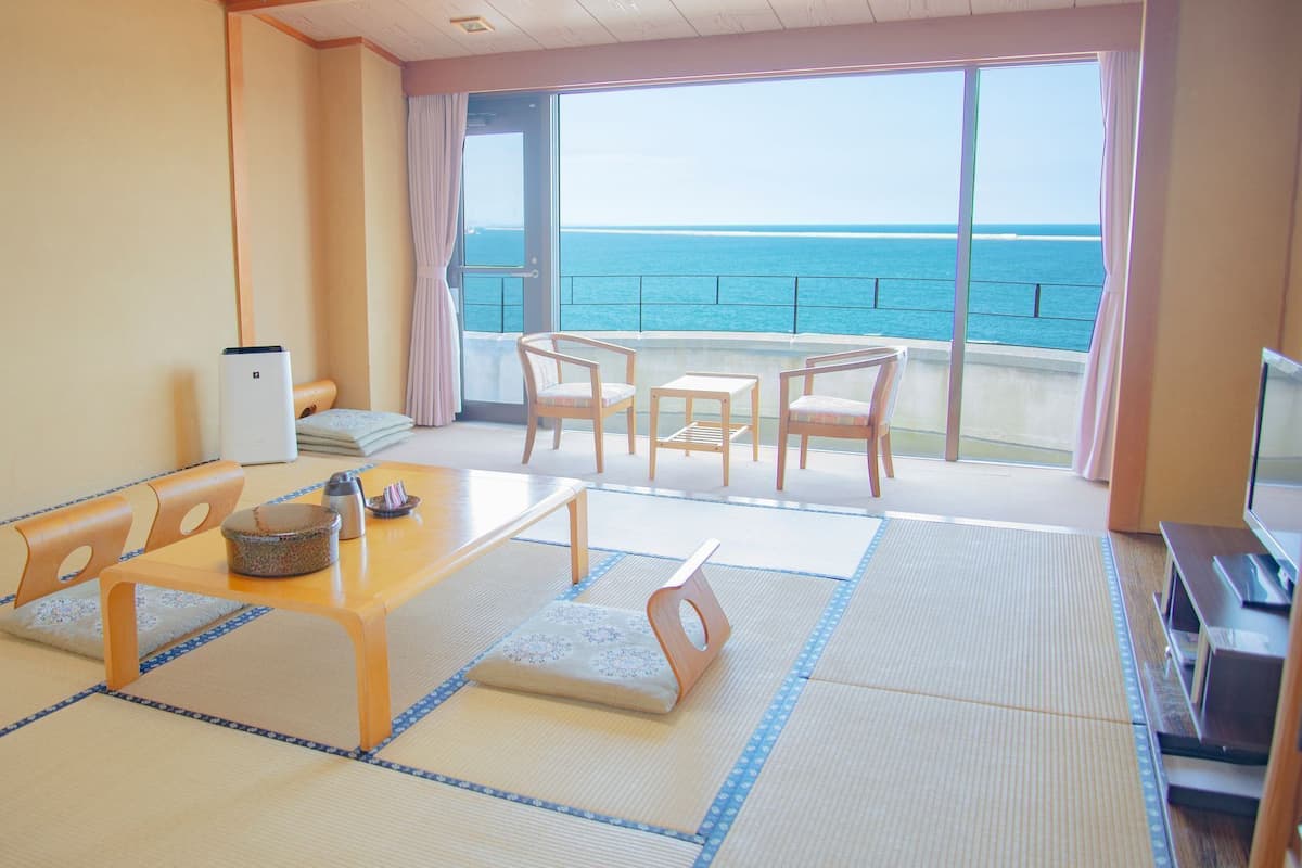A room with tatami flooring, a wooden table, chairs, a TV, and a view of the sea.