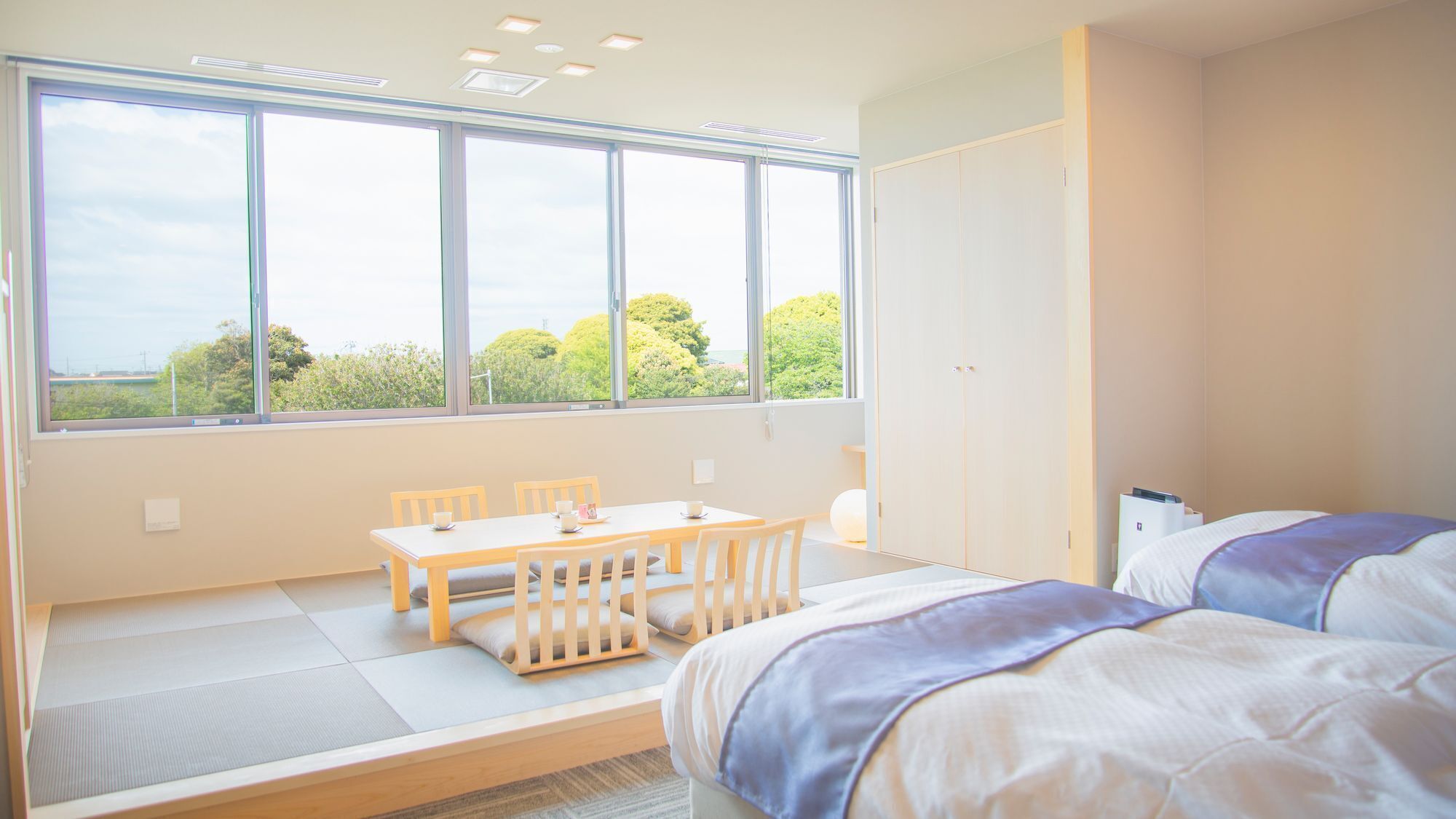 A modern hotel room with a large window, a dining table, two beds, and a wardrobe.
