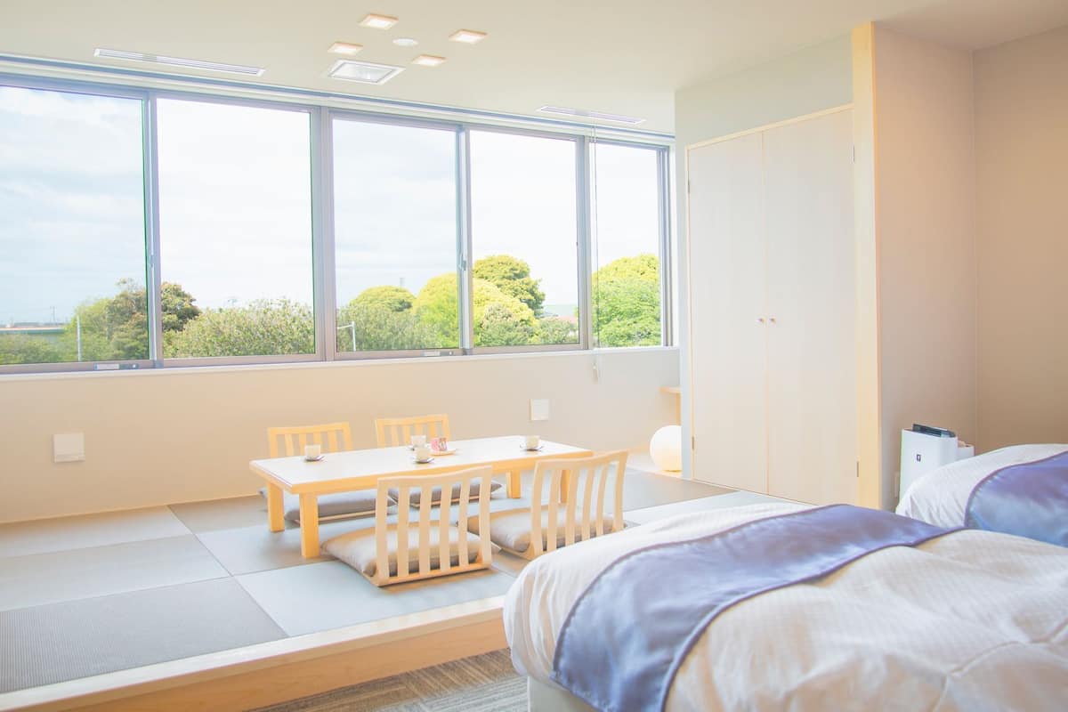 A modern hotel room with a large window, a dining table, two beds, and a wardrobe.