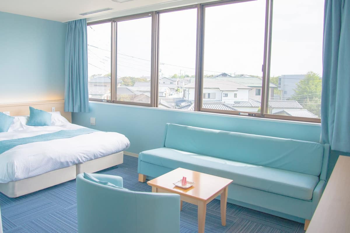 A hotel room with a bed, a sofa, a small table, and a window with a view of buildings.