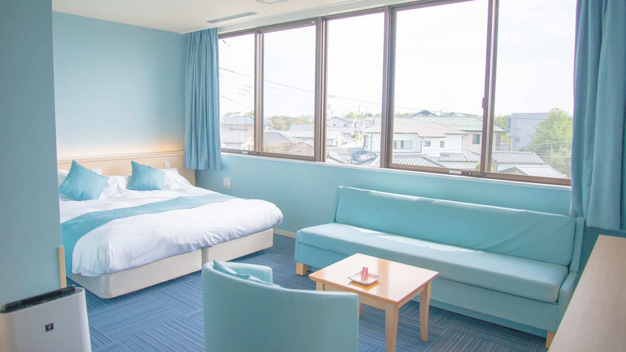 A hotel room with a bed, a sofa, a small table, and a window with a view of buildings.