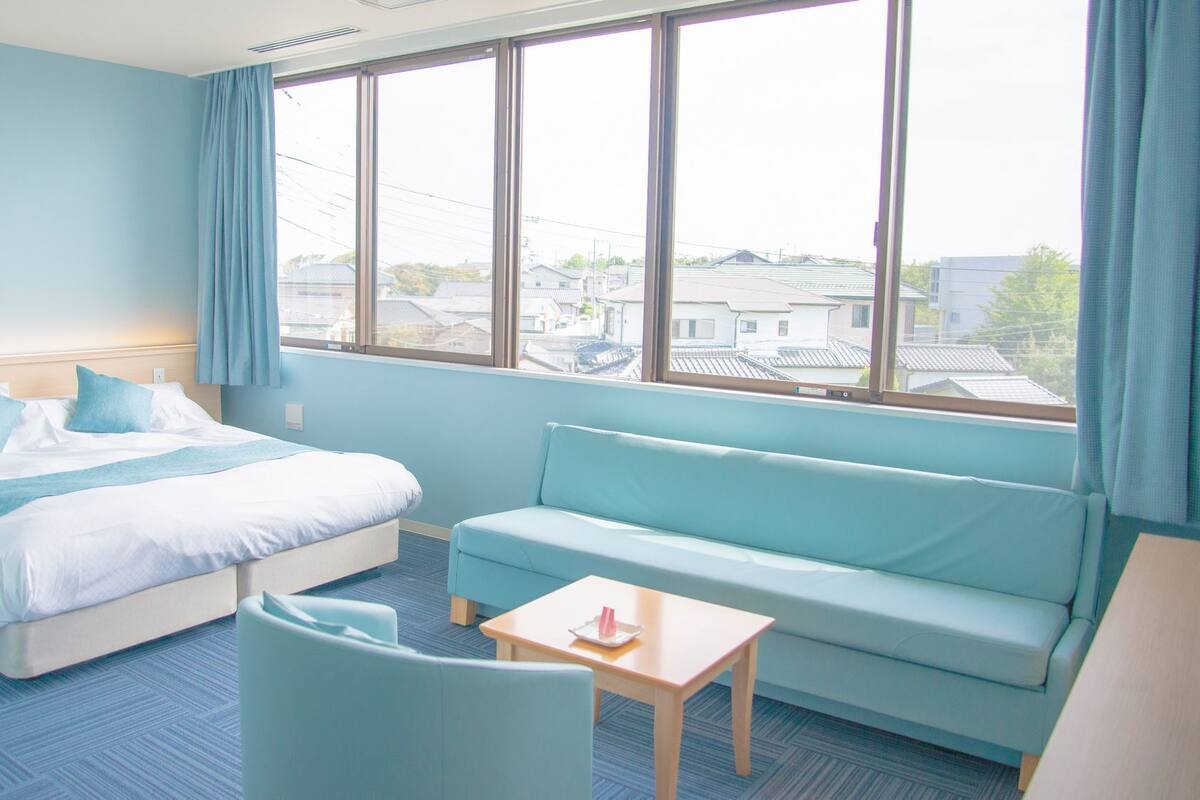 A hotel room with a bed, a sofa, a small table, and a window with a view of buildings.