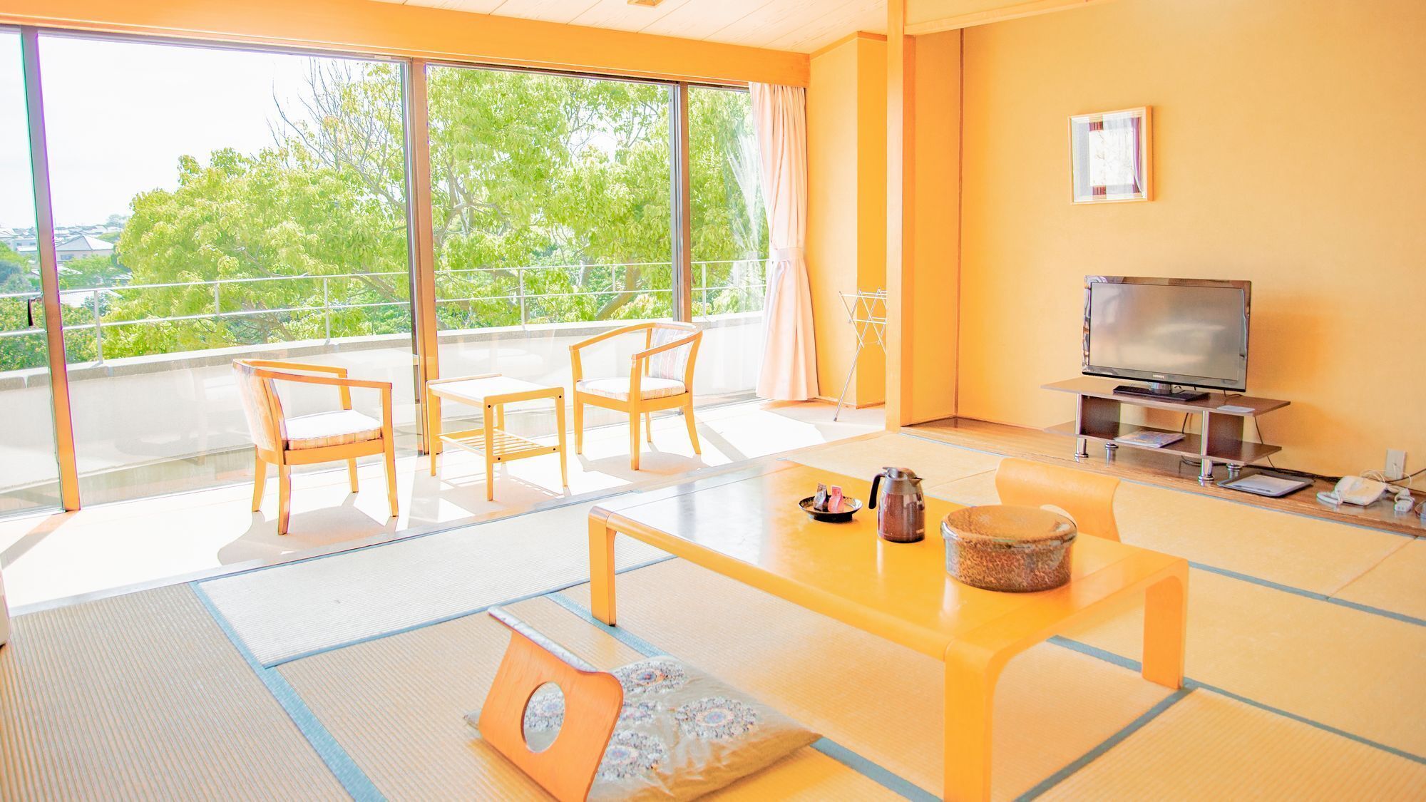 A modern living room with a TV, sofa, and chairs, large windows with a view of trees, and tatami flooring.