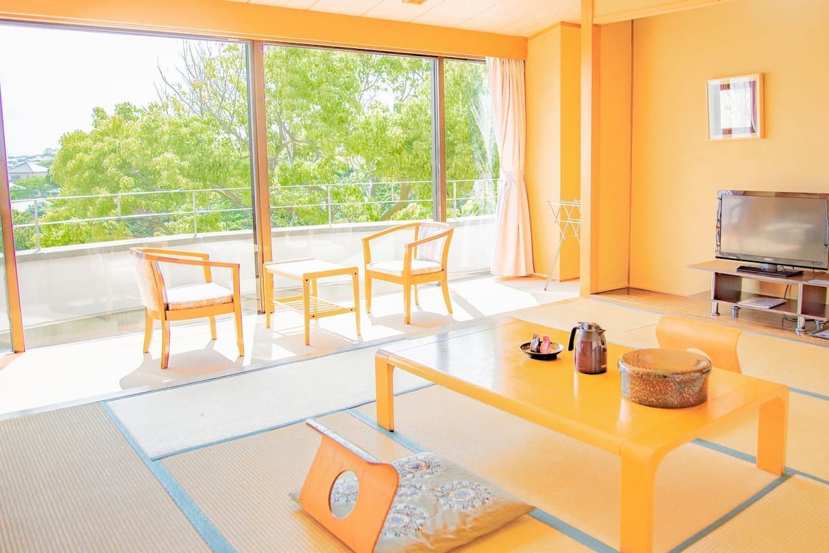 A modern living room with a TV, sofa, and chairs, large windows with a view of trees, and tatami flooring.
