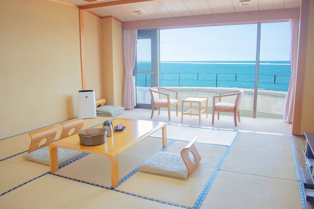 A room with tatami flooring, a low table with cushions, a TV, and a view of the sea.