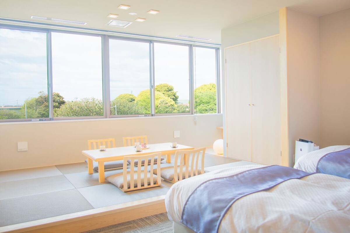 A hotel room with a large window, a dining table, two beds, and a wardrobe.