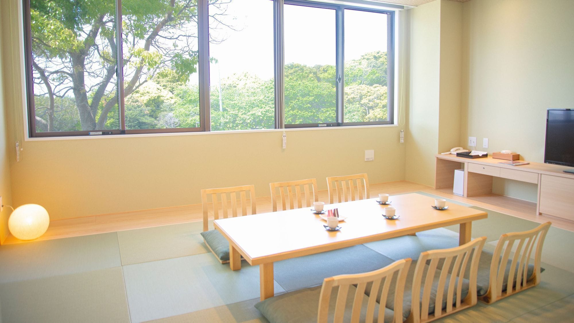 A room with a large window, a wooden table with chairs, a small desk with a computer, and a TV.