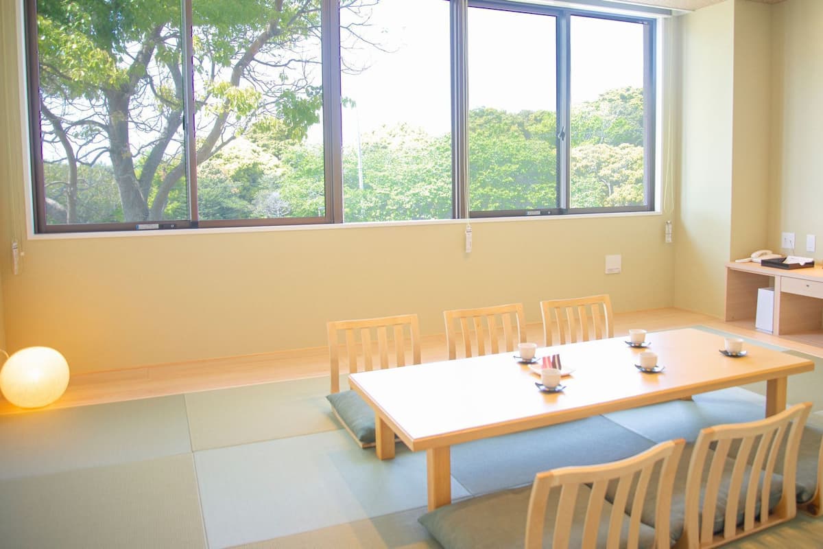 A room with a large window, a wooden table with chairs, a small desk with a computer, and a TV.