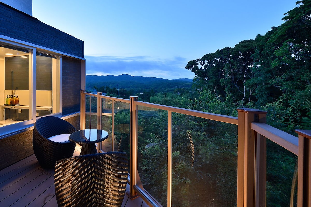 A balcony with wicker furniture, a glass door leading to a bathroom, and a view of a forested landscape.