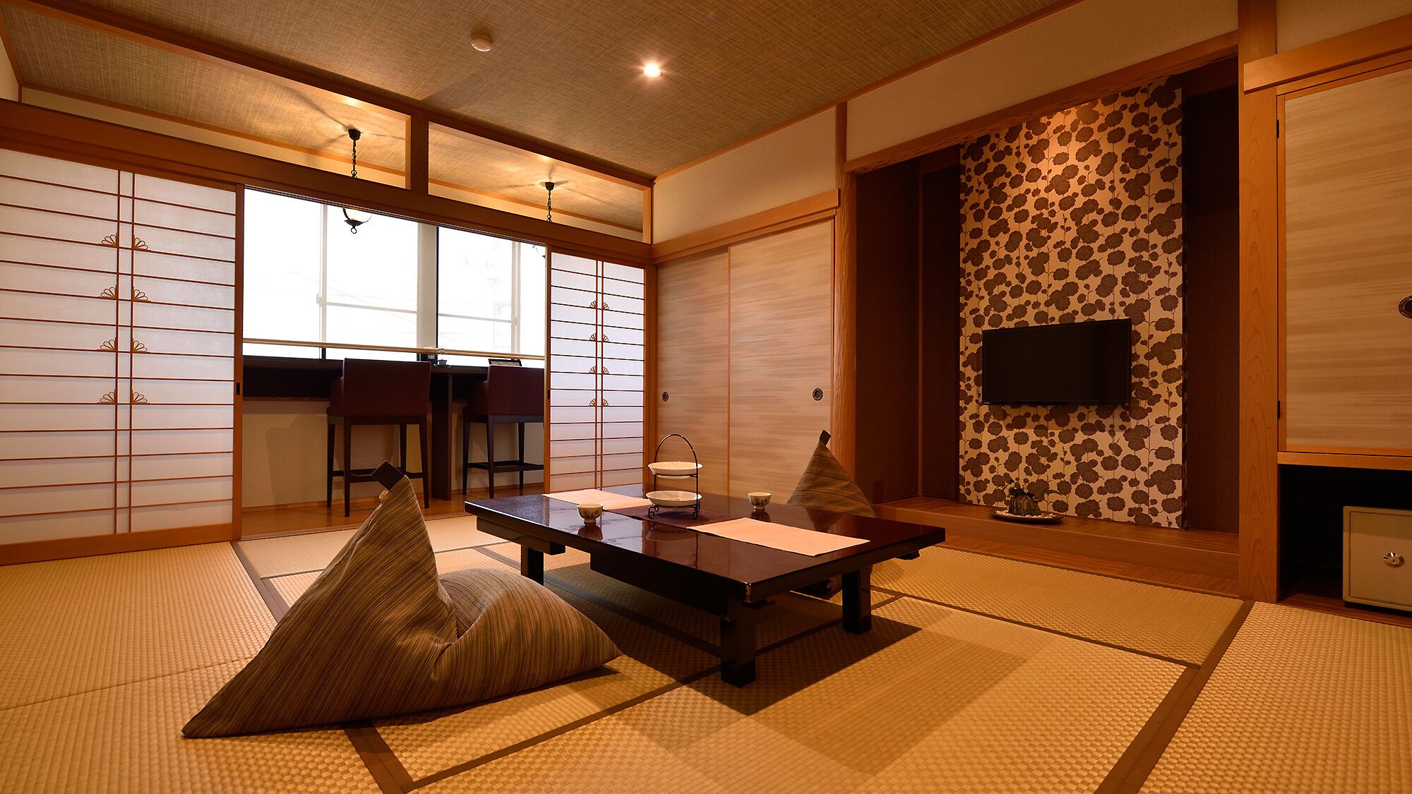 Um quarto japonês tradicional com tatames, uma mesa baixa com almofadas, portas deslizantes e uma televisão fixada na parede.