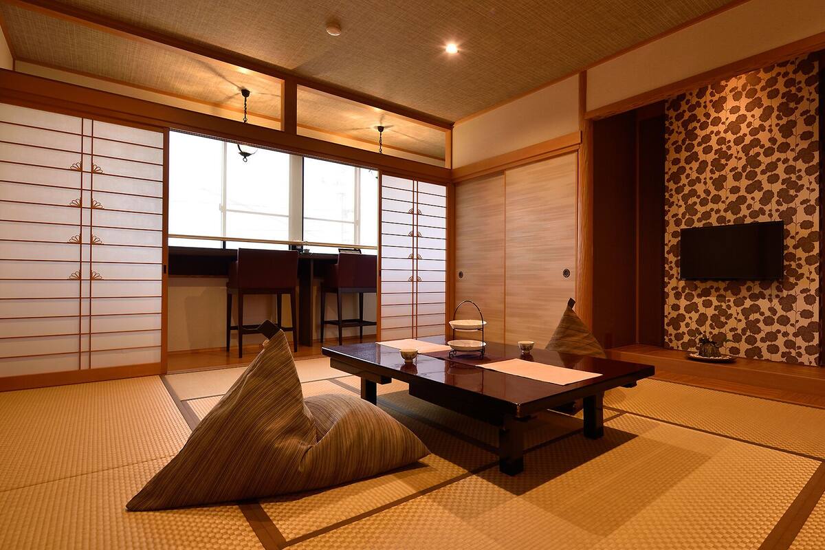 A traditional Japanese room with tatami flooring, a low table with cushions, sliding doors, and a television mounted on the wall.