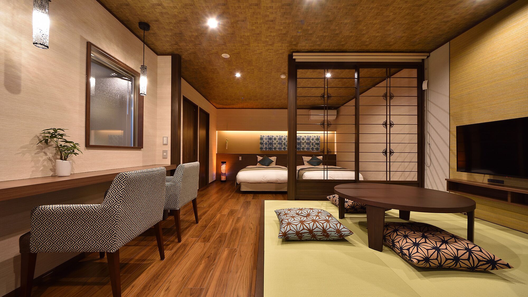 Quarto de hotel com piso de madeira, cama com cabeceira branca, televisão na parede, escrivaninha com cadeira e porta deslizante.